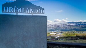 A sign reading "HRÍMLAND 10" with a mountainous background and a view of a landscape with buildings and a road. - Hrimland (Akureyri)