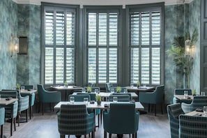 A restaurant with a modern interior, featuring a central dining table surrounded by chairs, large windows with blinds, and decorative wall sconces. - Lincombe Hall Hotel (Torquay)
