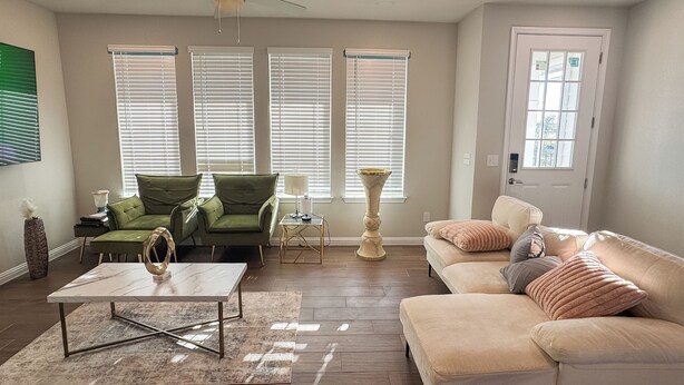 Ein Wohnzimmer mit einem beigen Sofa, einem grünen Sessel, einem Couchtisch und einer Stehlampe. Drei Fenster mit Jalousien, ein Deckenventilator und eine Tür mit Fenster sind vorhanden.