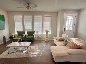 A living room with a beige sofa, a green armchair, a coffee table, and a floor lamp. There are three windows with blinds, a ceiling fan, and a door with a window. - Centrally located, beautiful new home (Austin)