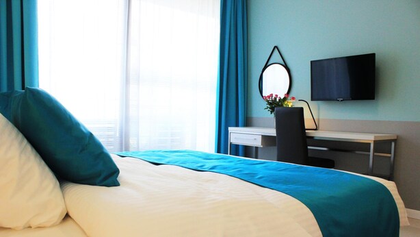A hotel room with a bed, a desk, a chair, a TV, and a window with curtains. - Jessapart Hotel - Triton Park (Warsaw)