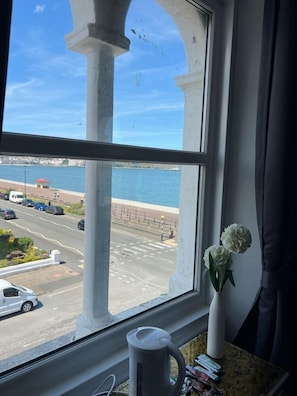 Une fenêtre donnant sur le bord de l’eau, un vase blanc rempli de fleurs et une tasse blanche posée sur une table.