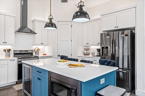 Eine moderne Küche mit weißen Schränken, einem Edelstahlkühlschrank, einem eingebauten Backofen und einer großen Kochinsel mit Obstschale und Schneidebrett.