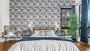 Un dormitorio con una cama grande, una silla, una mesita de noche, una planta en maceta y una ventana con cortinas.