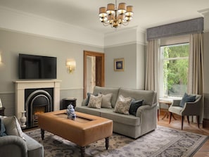 A living room with a grey sofa, a coffee table, a fireplace, a television, and a large window with curtains. - THE Marcliffe Hotel AND SPA (Aberdeen)