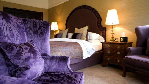 Une chambre d’hôtel avec un grand lit, deux lampes de chevet, une table de chevet en bois et un fauteuil violet.