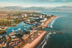 Vista aerea di una località balneare con un porto turistico, spiaggia e montagne sullo sfondo.