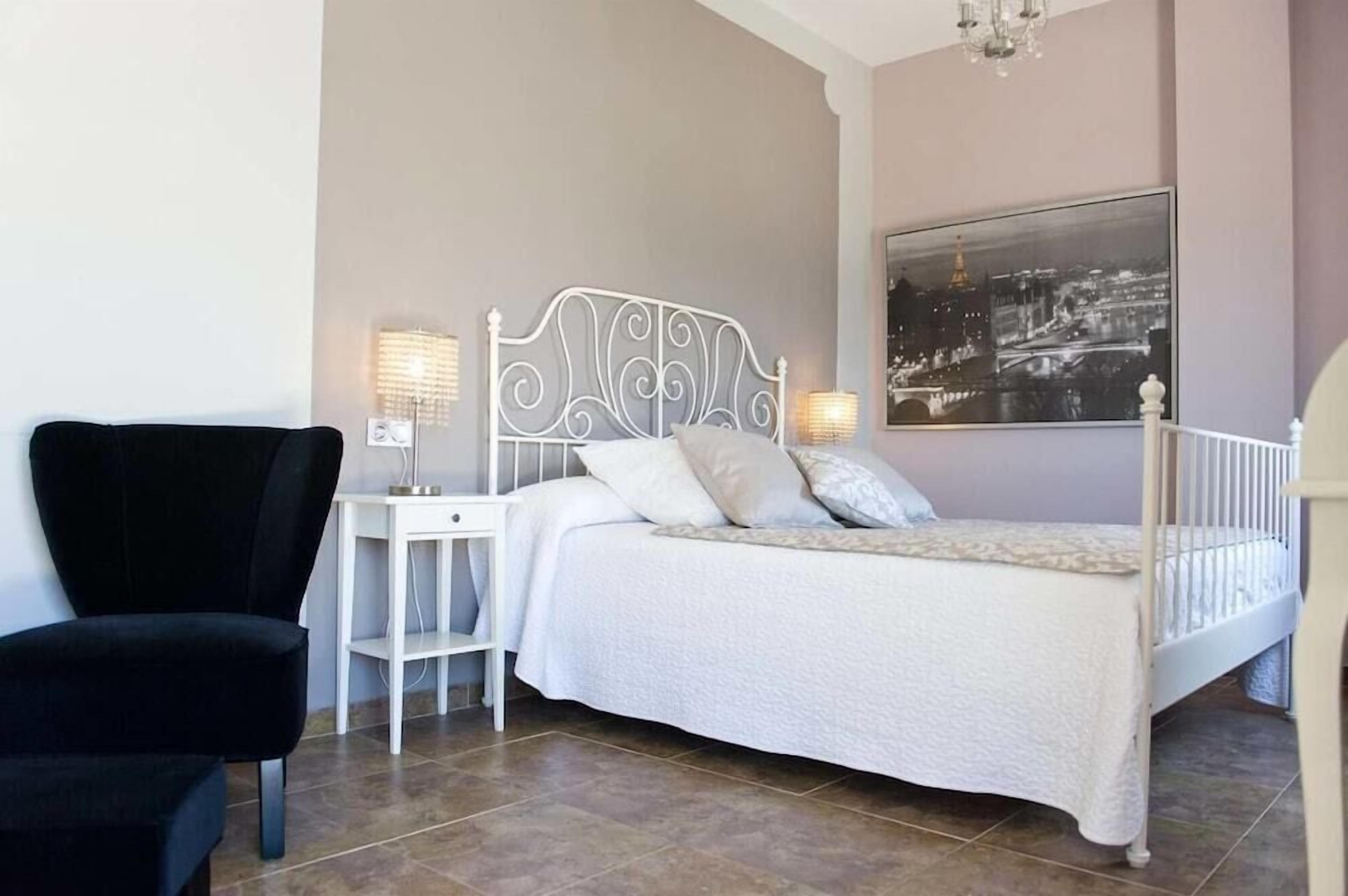 Un dormitorio ordenado con cama, mesitas de noche, una silla y un cuadro grande con una vista urbana.