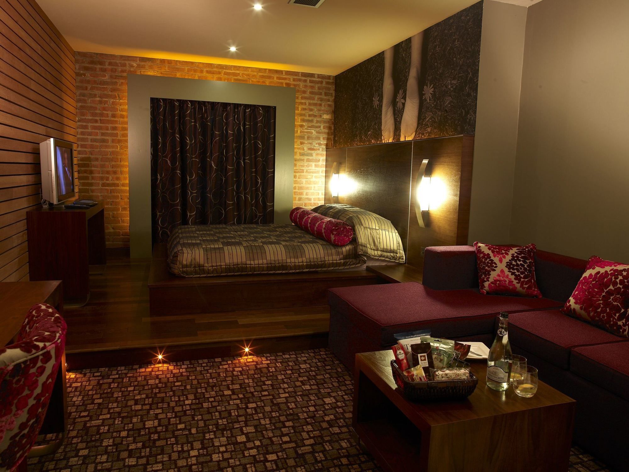 A modern hotel room with a brick accent wall, a large bed, a sofa, and a small table with a bottle and glasses.