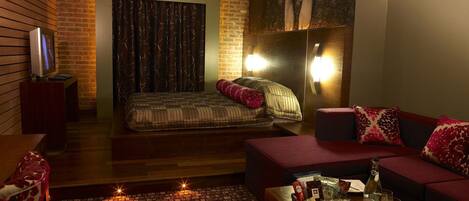 A modern hotel room with a brick accent wall, a large bed, a sofa, and a small table with a bottle and glasses.