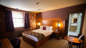 A hotel room with a wooden bed, a small table, a chair, a window with curtains, and a bathroom visible through an open door. - The Crown Hotel (Doncaster)