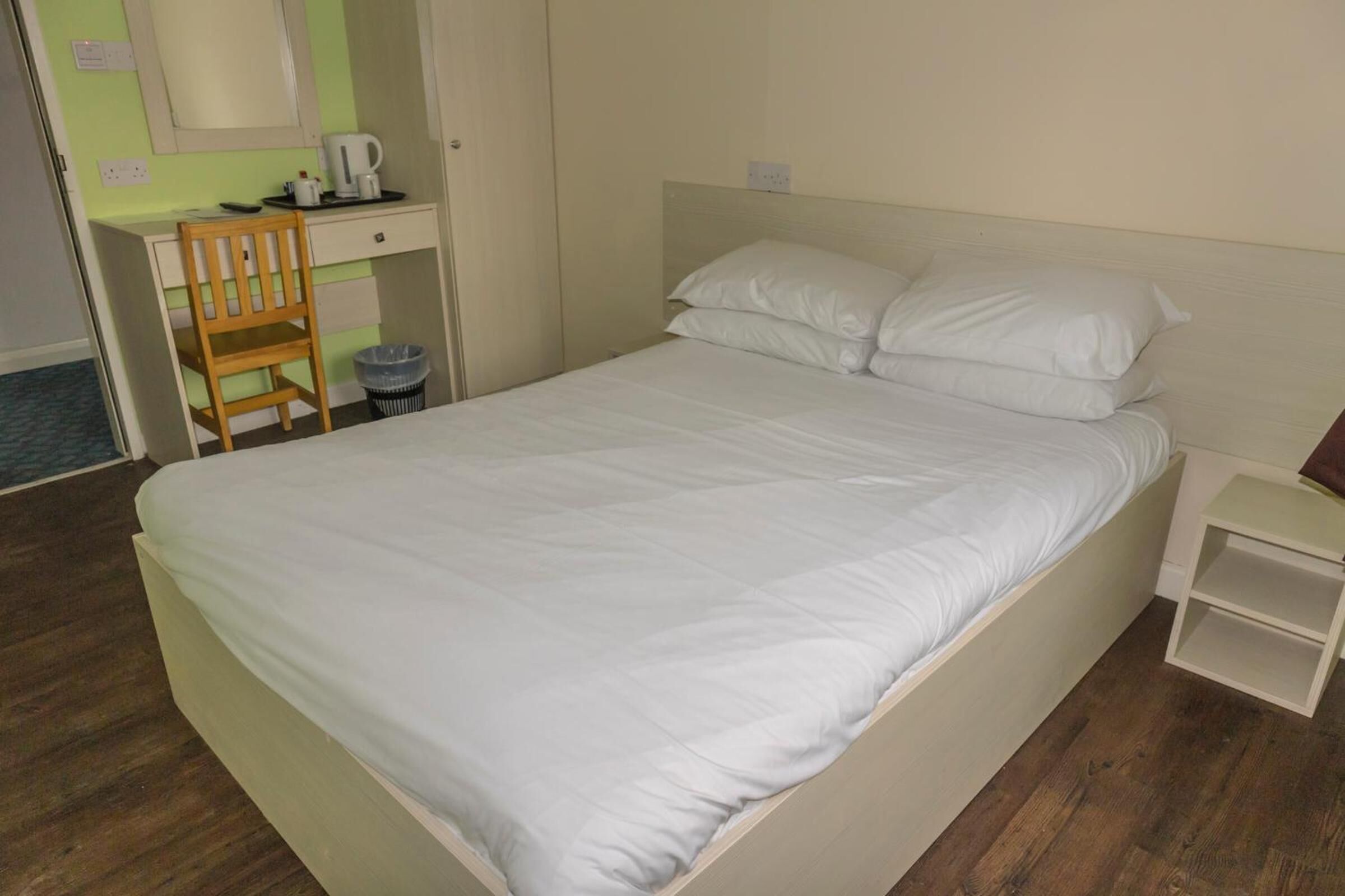 A bed with white bedding, a wooden chair, a desk with a coffee maker, and a small bedside table.