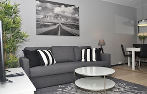 A modern living room with a grey sofa, two round tables, a dining area with a white table and black chairs, and a large black and white road photograph on the wall. - Forenom Apartments Carl Berners Plass (Oslo)