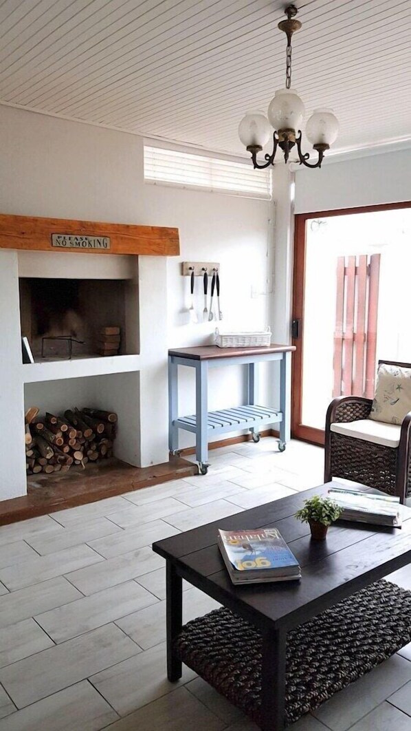 A living room with a fireplace, a coffee table with magazines, a wicker chair, and a hanging light fixture. - Normandie On Sea (Hermanus)