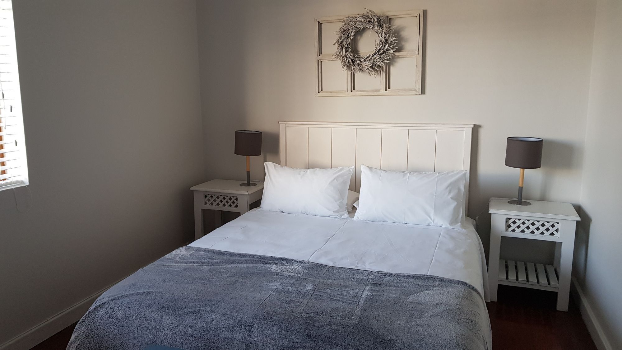 A bedroom with a bed, two bedside tables, a nightstand, a wall-mounted light fixture, and a decorative wreath.