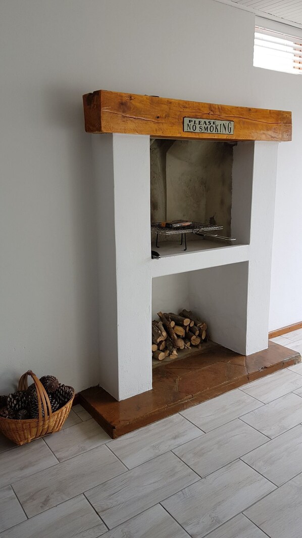 A fireplace with a 'No Smoking' sign above it, a basket of pinecones, and a stack of firewood. - Normandie On Sea (Hermanus)