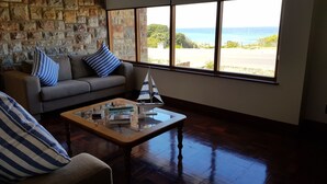 A living room with a stone wall, a sofa with striped pillows, a coffee table with books and a model sailboat, and a view of the ocean through large windows. - Normandie On Sea (Hermanus)