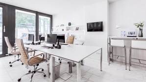 Un espacio de oficina moderno con un escritorio grande de color blanco, sillas ergonómicas, un televisor de pantalla plana y una pequeña zona de bar con taburetes.