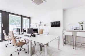 A modern office space with a large white desk, ergonomic chairs, a flat-screen TV, and a small bar area with stools. - Visionapartments Frankfurt Gutleutstraße (Frankfurt)