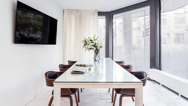 Una sala de reuniones moderna con una mesa de vidrio, seis sillas, un florero con flores, un cuadro en la pared y ventanas grandes con vistas a la ciudad.