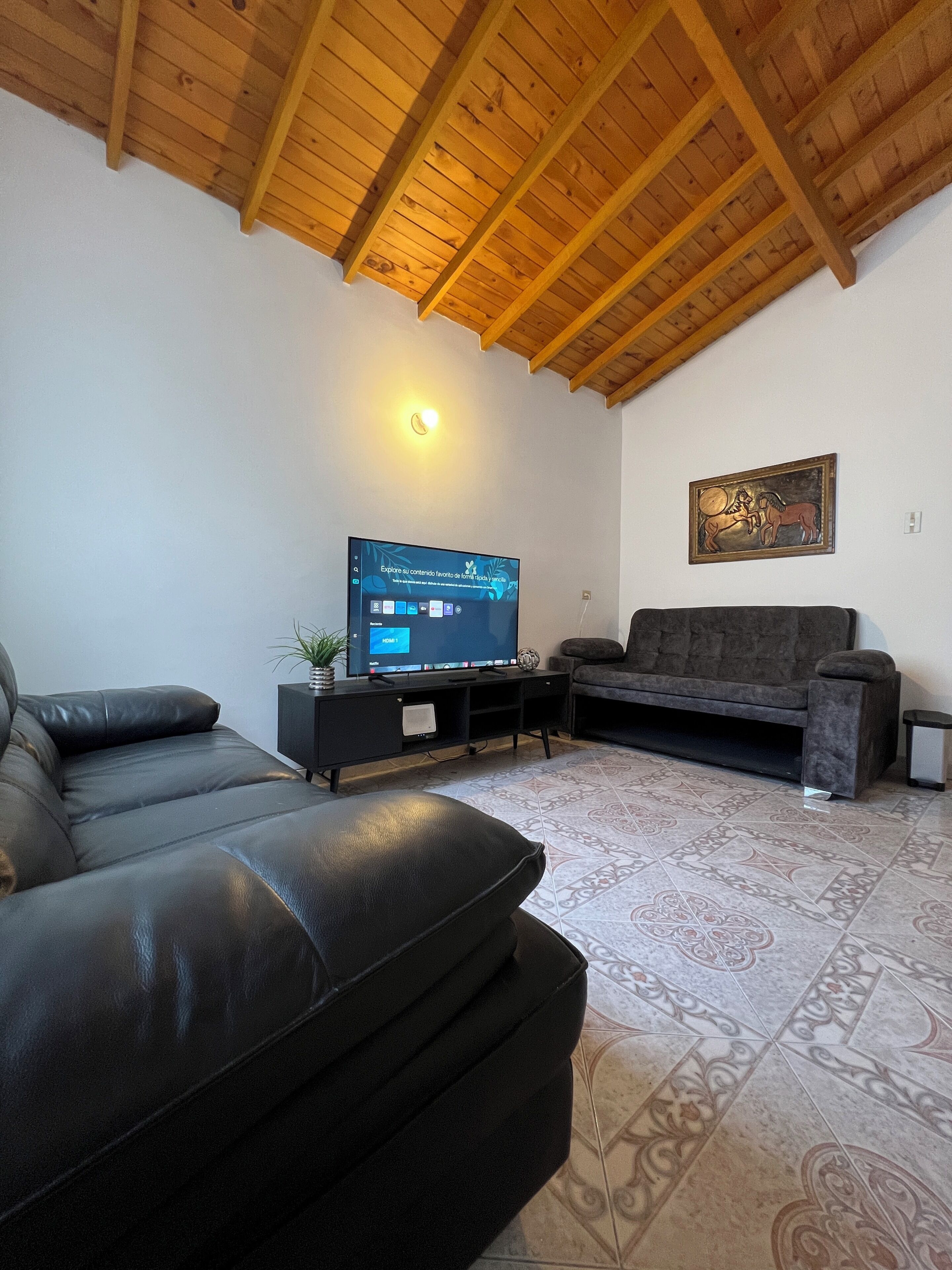 Una sala de estar con techo de madera, un sofá de cuero negro, un sofá gris, un televisor sobre un soporte negro y un piso con diseño.