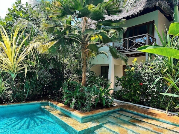 A pool area with a thatched roof structure, surrounded by lush tropical plants and a building with a balcony. - Aluna Beach Lodge (Nungwi)