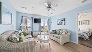 A room with wicker furniture, a ceiling fan, a TV, and a view through a glass door. - The East Beach Classic | Ground Floor Convenience (Gulf Shores)