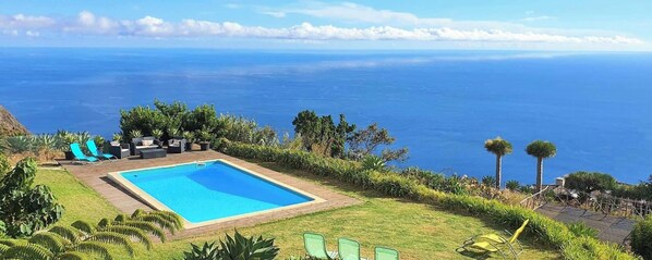 A rectangular pool with a wooden deck, surrounded by green chairs and potted plants, overlooking a vast ocean. - Quinta São Lourenço - Casa Frente | Sunset - Pool - Seaview (FAJÃ DA OVELHA)