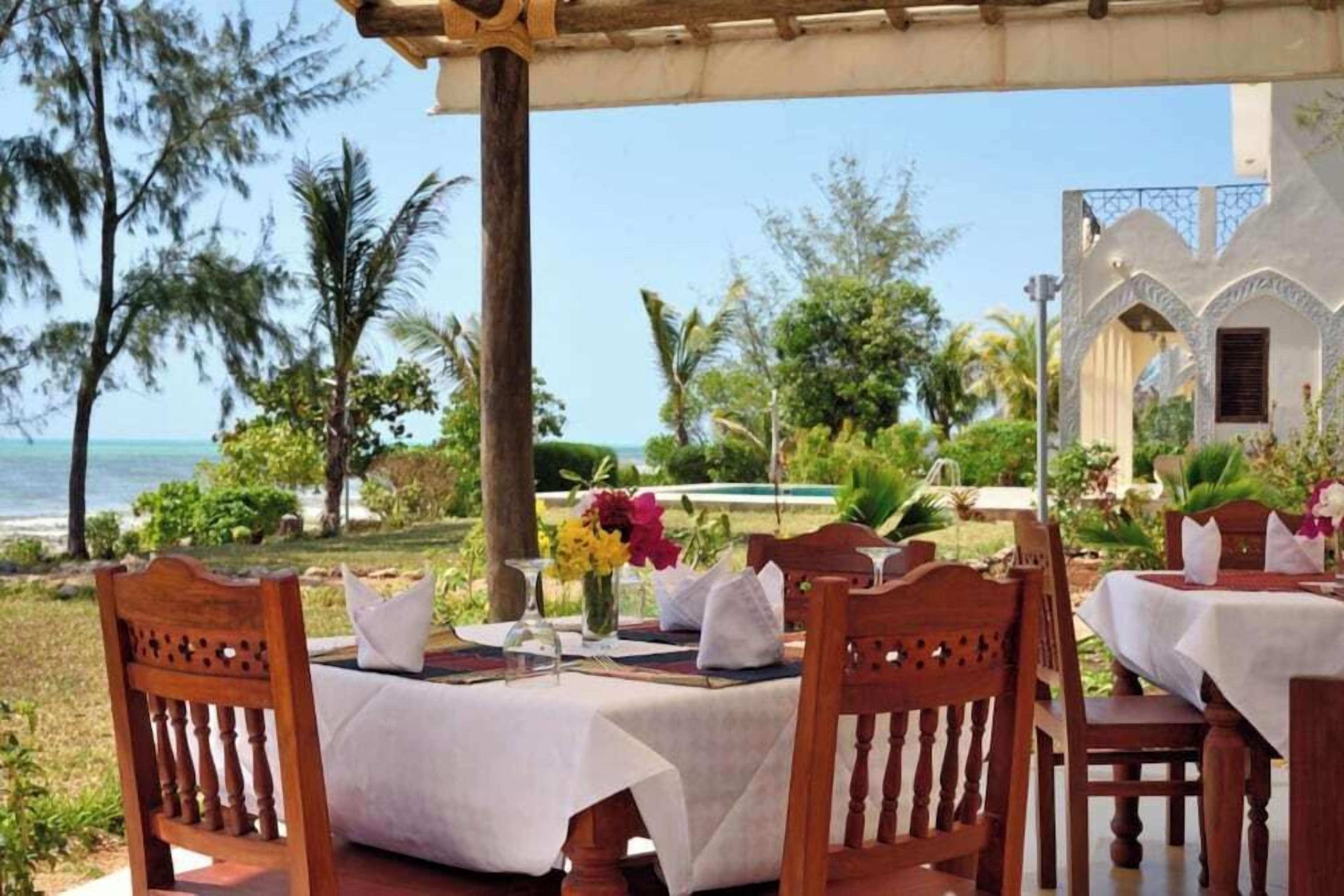Un espace extérieur abrité, aménagé pour les repas, avec des chaises et des tables en bois, une vue sur l’océan et une piscine en arrière-plan.