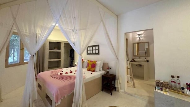 A bedroom with a canopy bed, a bedside table, a small nightstand, a mirror, and a bathroom with a sink and toiletries. - Sharazad Villa (Jambiani)