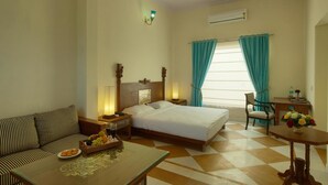 A hotel room with a bed, a sofa, a table with fruit and glasses, a chair, a desk with a vase of flowers, and a window with curtains. - Aaram Baagh Agra On Pachar Group (Pushkar)