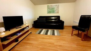 A living room with a black leather sofa, a wooden entertainment unit with a TV, a remote, and a lamp. There's a striped rug on the wooden floor. - Skálatjörn Homestay (Selfoss)