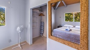 Ein Schlafzimmer mit einem Bett, einem Nachttisch, einem Fenster mit Blick auf Palmen und einem Ventilator in der Ecke.