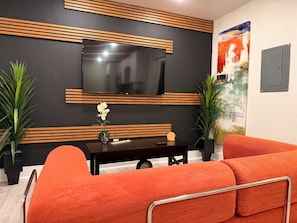 A modern living room with a flat-screen TV mounted on a dark wall, a wooden accent wall, a black coffee table, orange sofas, and two potted plants. - Downtown Dallas Hideaway | Free Parking | 4 Guests (Dallas)