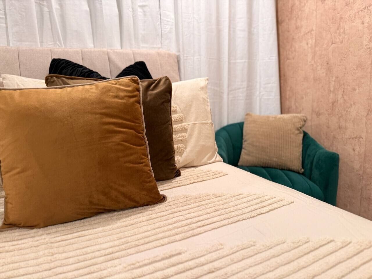 A bed with a textured white cover, accompanied by three pillows in varying shades of brown and beige.