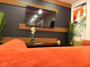 Ein modernes Wohnzimmer mit einem flachbildfernseher an einer dunklen Wand, Holzlamellen, einem kleinen Tisch mit einer Blumenvase und einem Gemälde.