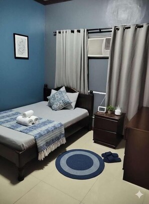 A bedroom with a bed, a bedside table with a lamp, a window with curtains, and a wall-mounted air conditioner. - Mactan-cebu Airport Budget Hotel (Lapu-Lapu)