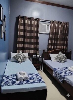 A hotel room with two single beds, each with a patterned bedspread and matching pillows. There is a small bedside table with a lamp and a clock. The room has a window with curtains, and the walls are painted blue. - Mactan-cebu Airport Budget Hotel (Lapu-Lapu)