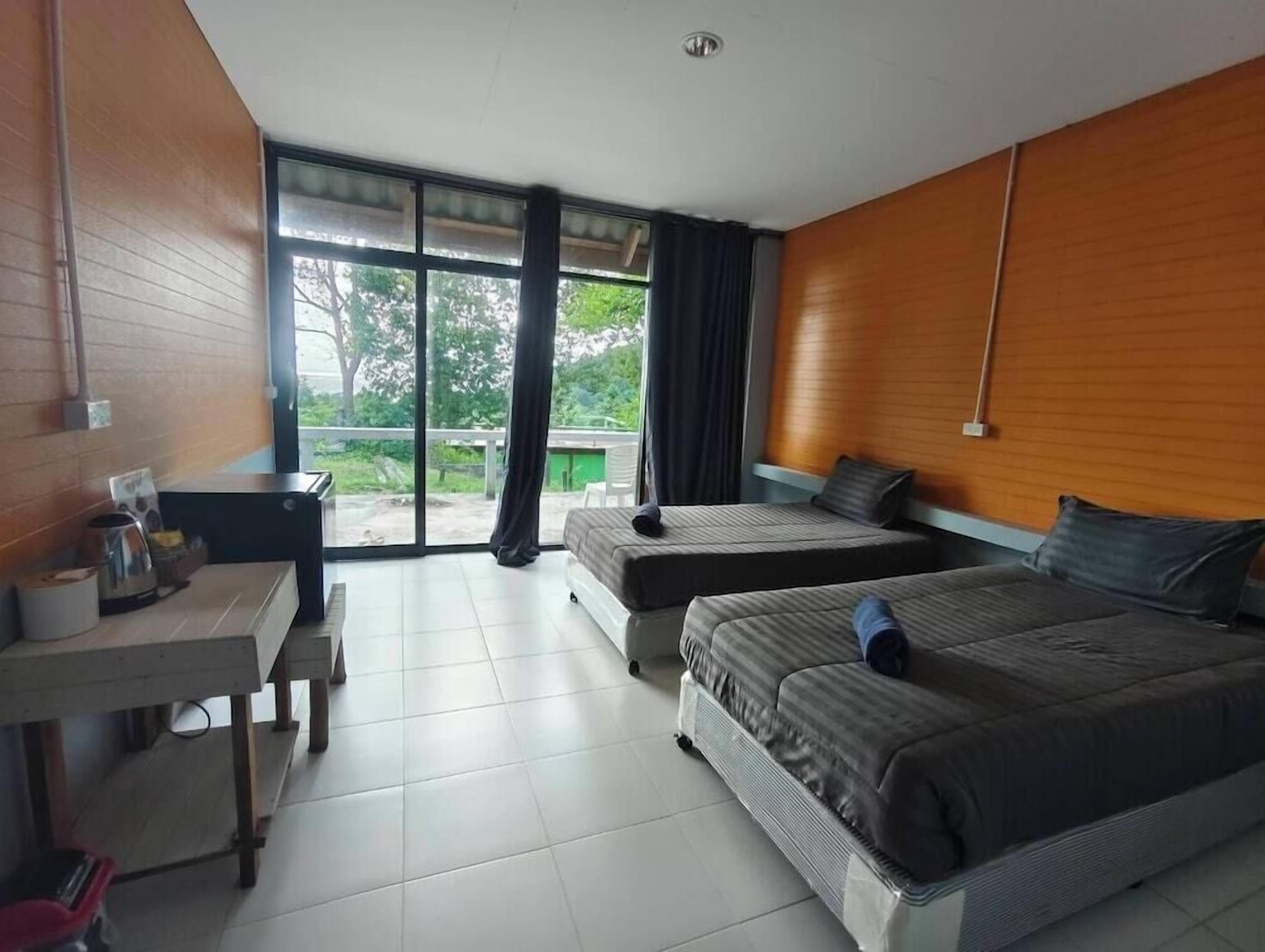 A hotel room with two beds, a desk, a chair, a window with curtains, and a view of greenery outside.