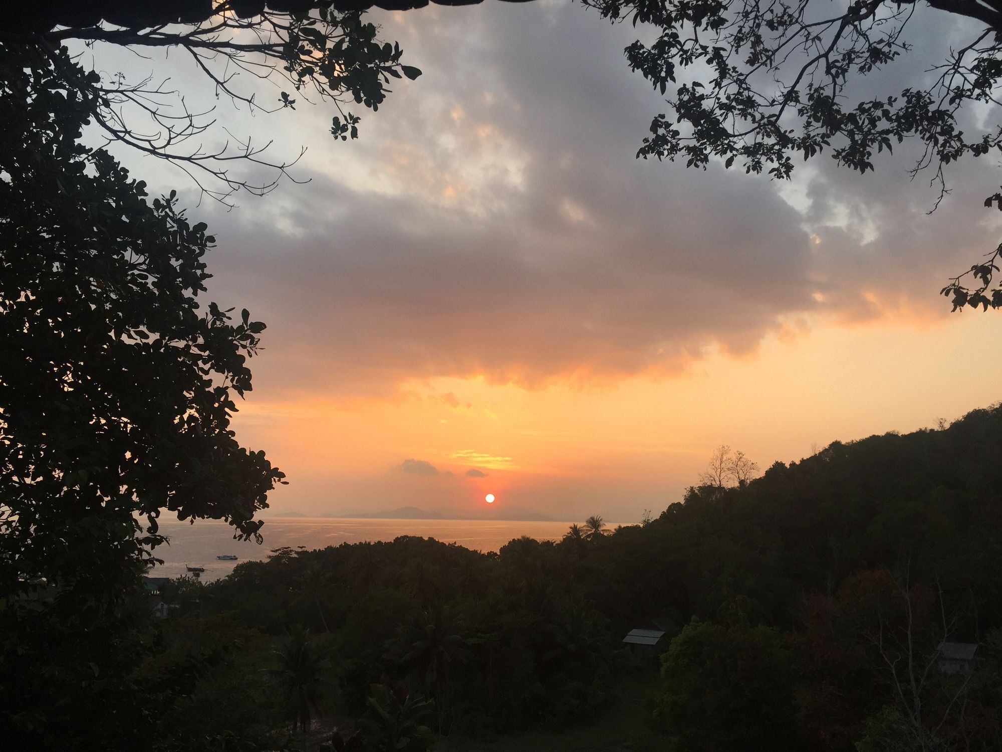 A sunset over a hilly landscape with dense foliage and a small building in the foreground.