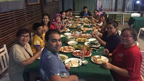 A group of people seated around a long table with various dishes, in a room with wooden paneling and framed pictures on the walls. - Banhalawee (Phang Nga)