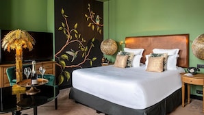 Hotelkamer met een groot bed, groene wanden, een muurschildering van vogels, een televisie en een tafel met een lamp en wijnglazen.