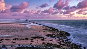Een strand met een rotsachtige kustlijn, een pier die in het water steekt, en een lucht in zonsondergangtinten.