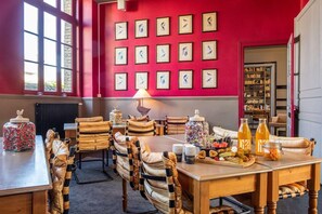 Un coin repas avec un mur rouge, des tableaux encadrés, une table dressée avec des assiettes, des verres et un plateau de fruits.
