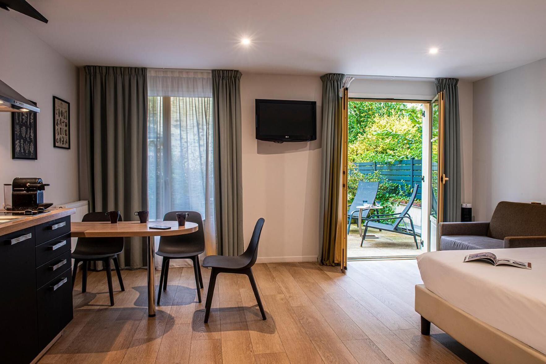 A modern hotel room with a wooden floor, a bed, a desk with a coffee machine, a TV, and a balcony with outdoor furniture.