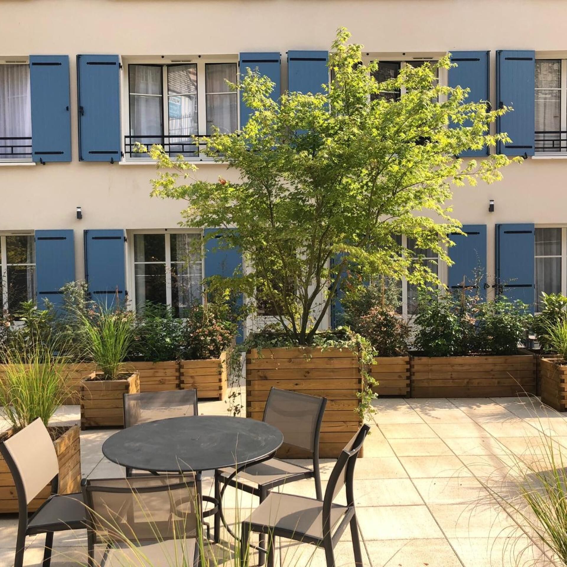 A courtyard with a round table and chairs, wooden planters, and a tree.