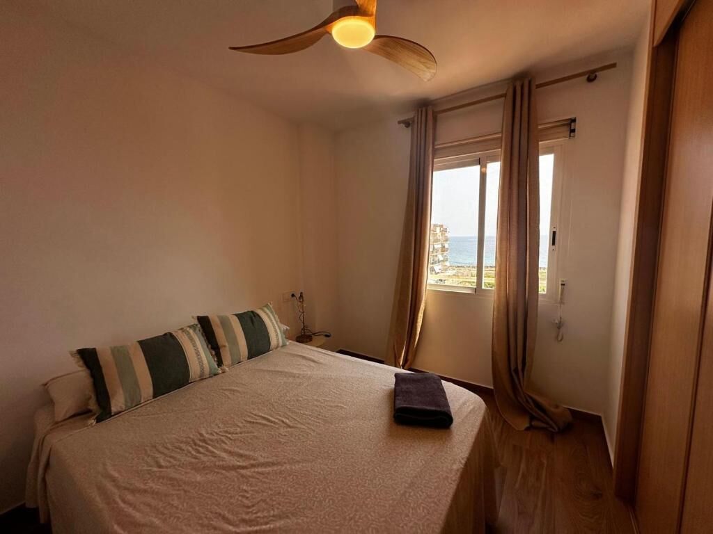 A bedroom with a bed, a ceiling fan, a window with curtains, and a view of the sea.