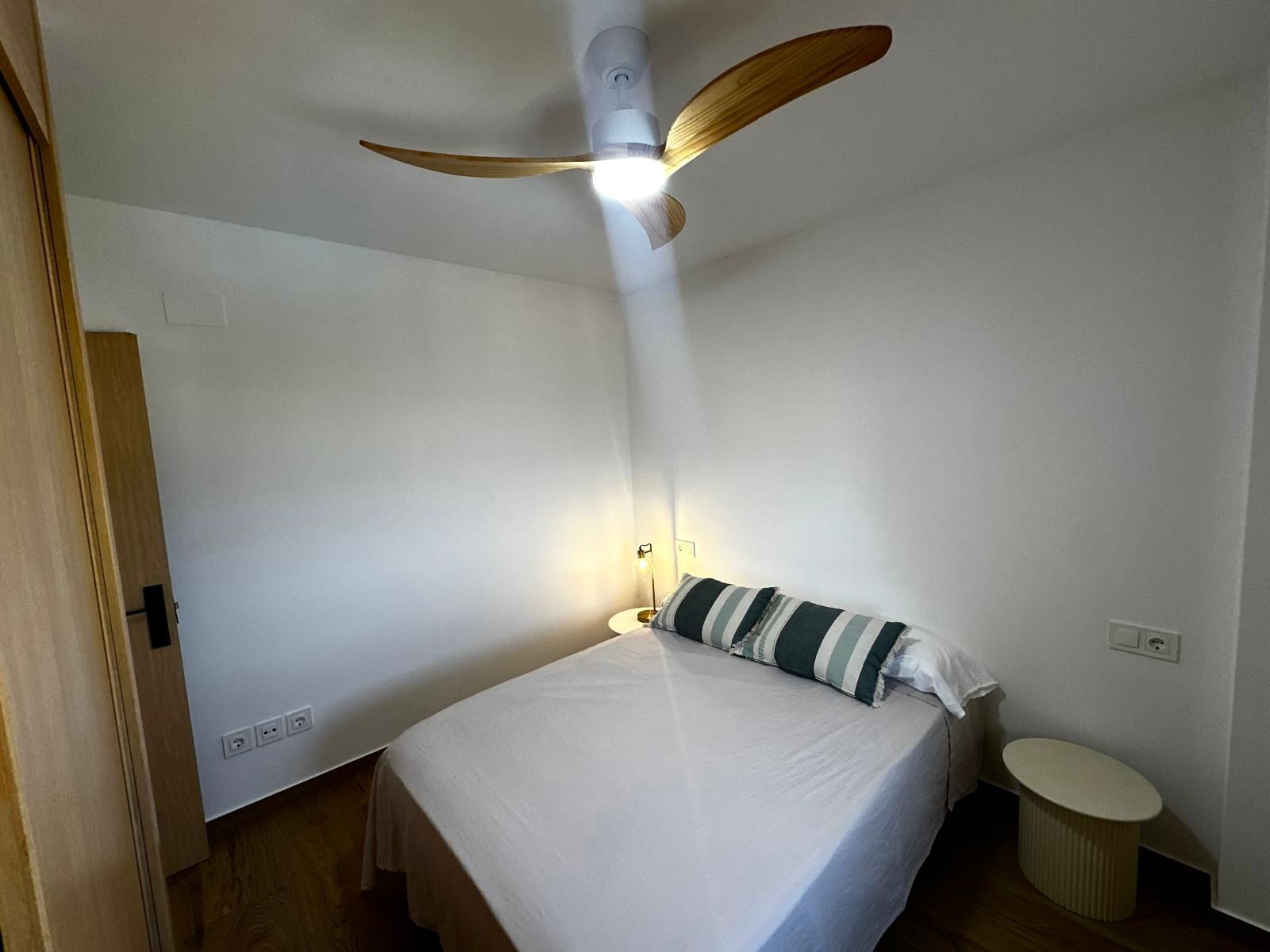 A single-bed room with a ceiling fan, bedside lamp, and a small round table.