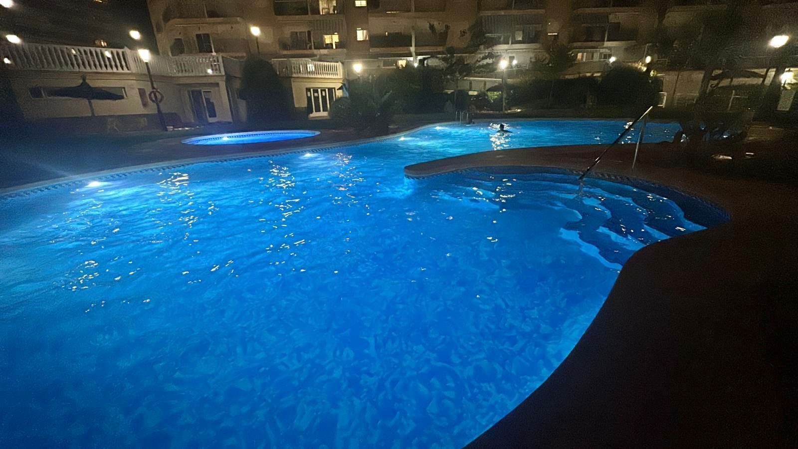 A swimming pool with a curved edge, surrounded by a building with balconies and lights.