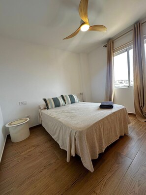 Un dormitorio con cama, ventilador de techo, ventana con cortinas y un pequeño cubo de basura.
